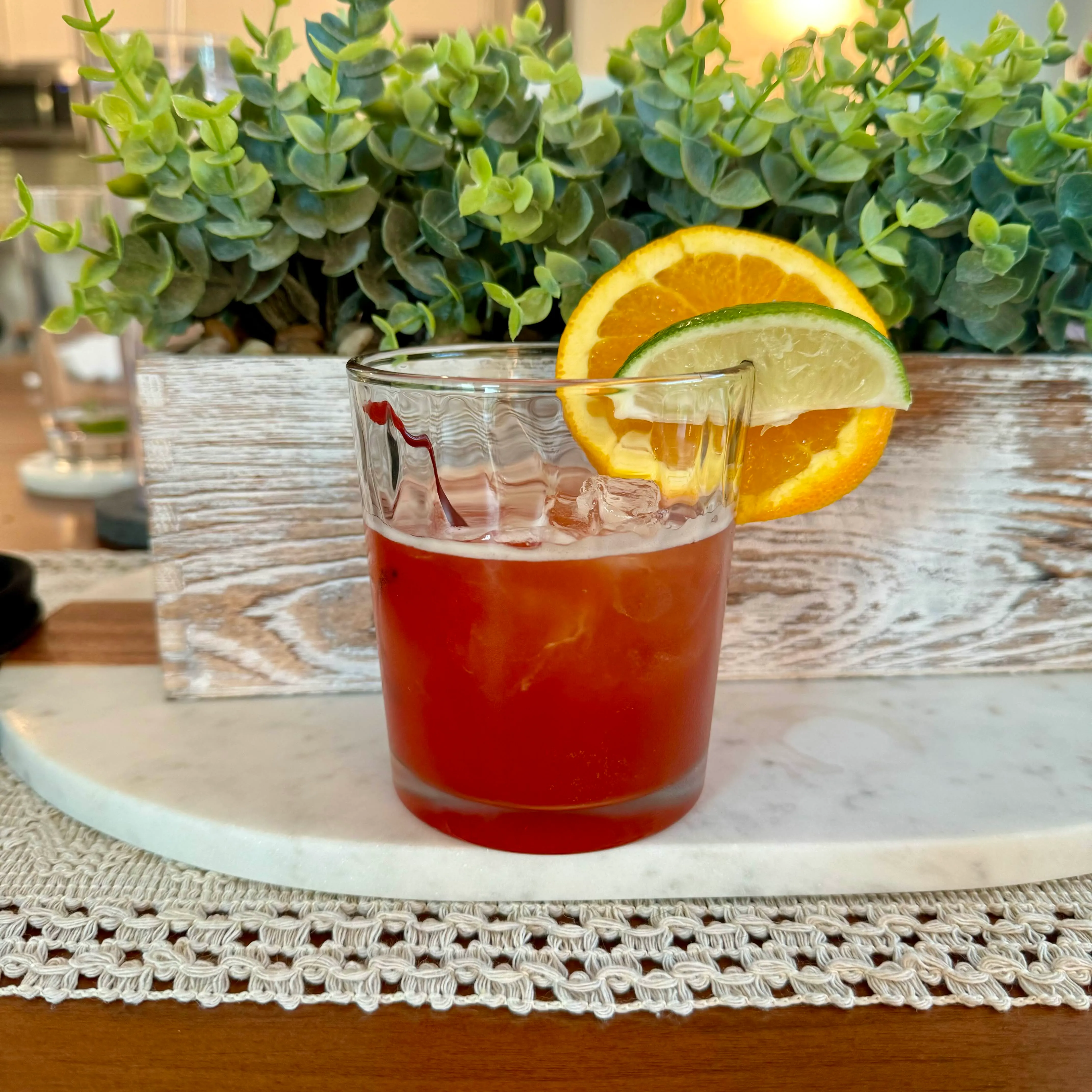 An amber cocktail over ice in a heavy-bottomed glass, garnished with a half-moon of orange and a wedge of lime.