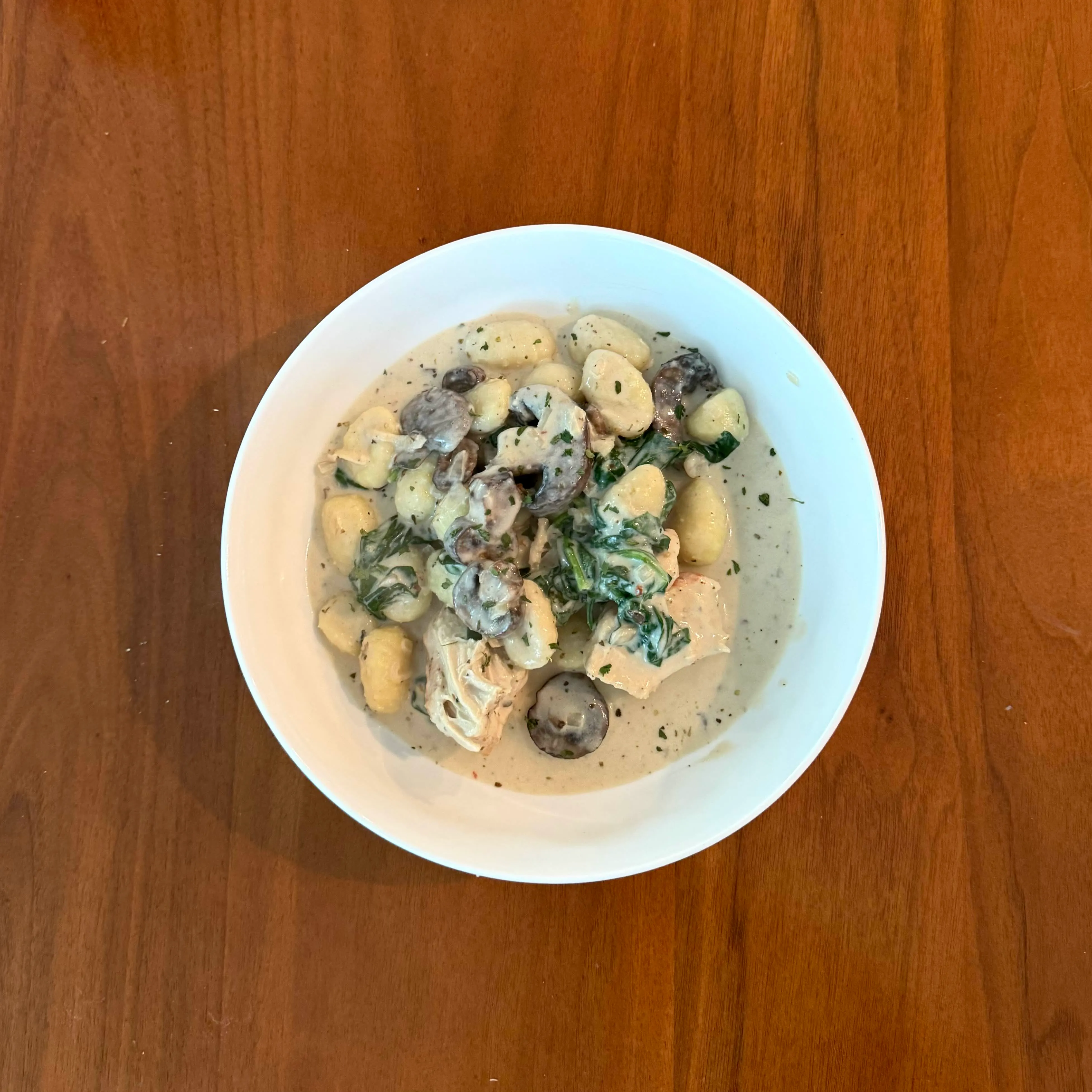 A white bowl of gnocchi with chicken, mushrooms, and spinach in a pale cream sauce, photographed from above on a warm wood table.
