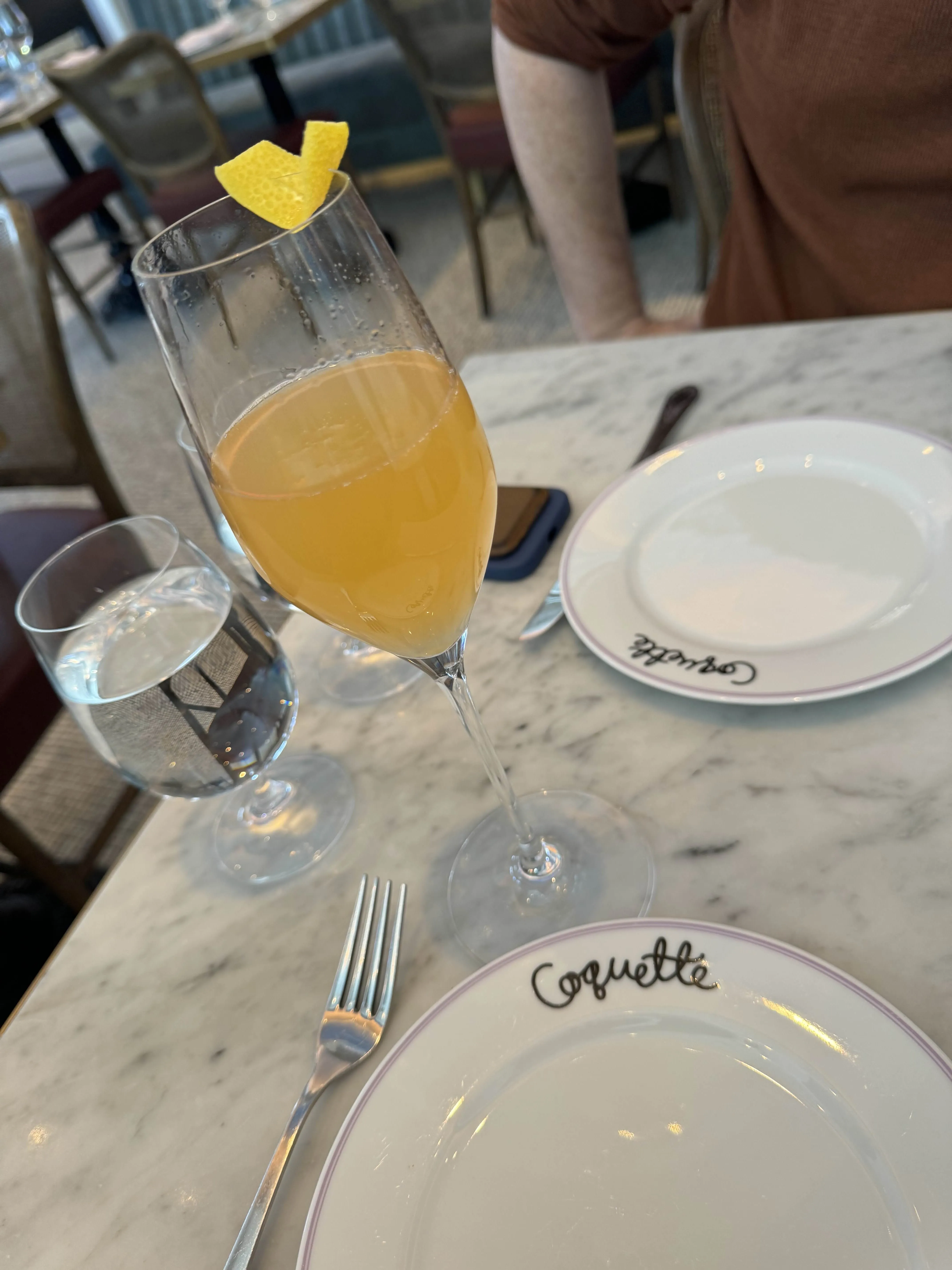 A champagne flute of pale mimosa garnished with a curl of lemon peel, set on a Coquette-printed plate.