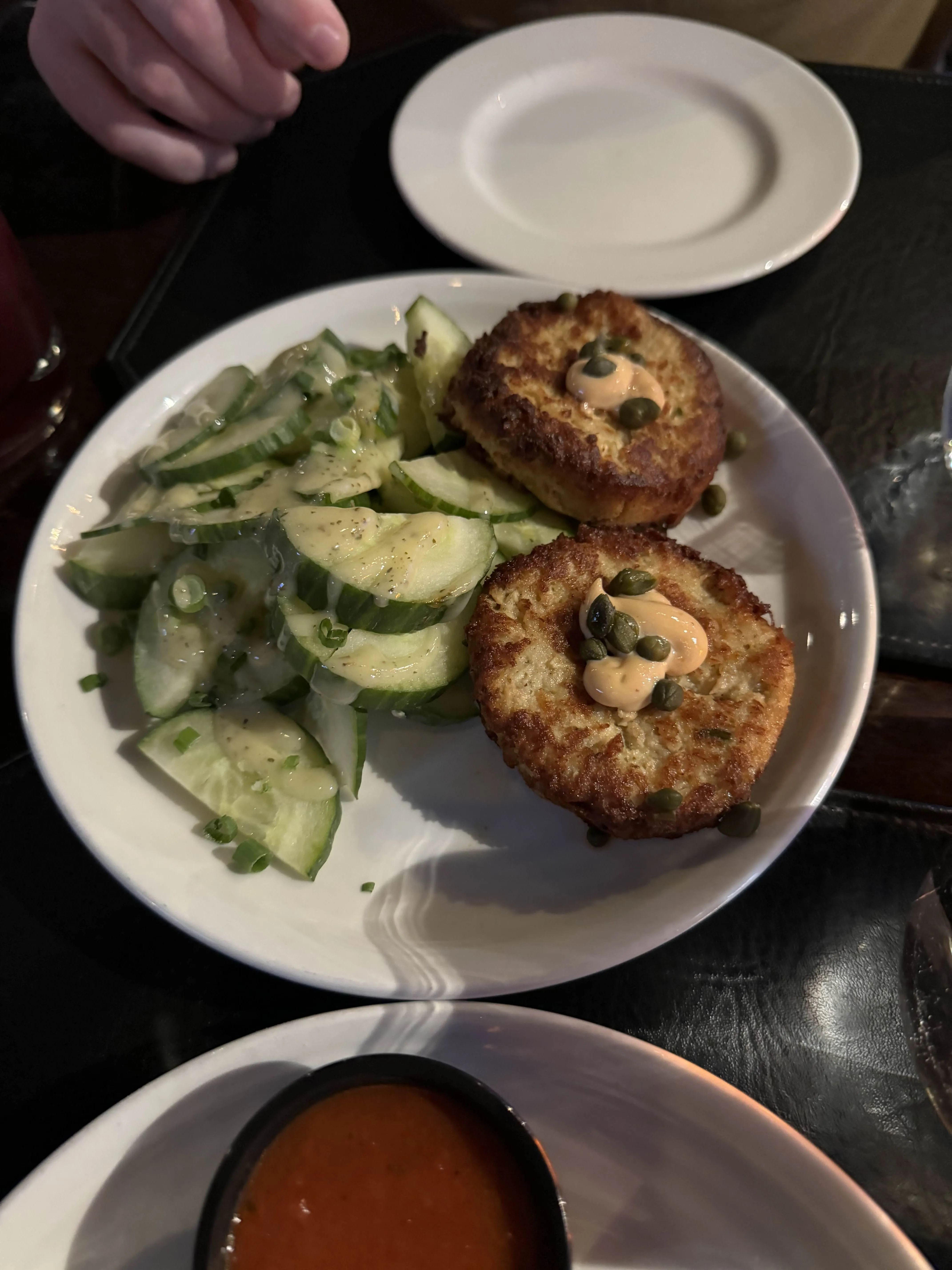Two golden-crusted crab cakes topped with a dot of aioli and capers, plated with a tangle of sliced cucumbers dressed with herbs.