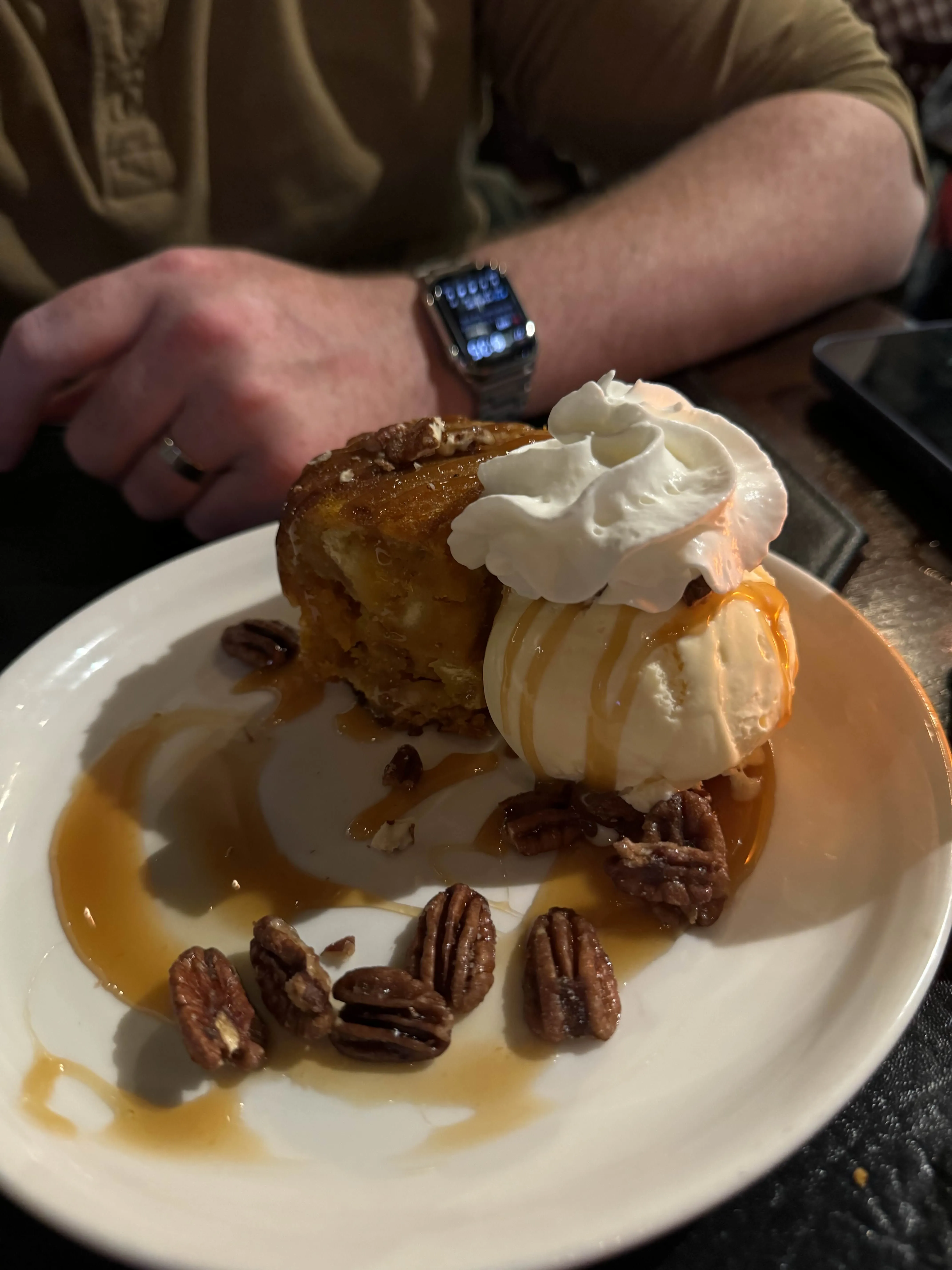 A round of warm bread pudding with a scoop of vanilla ice cream, caramel drizzle, and a scatter of candied pecans.