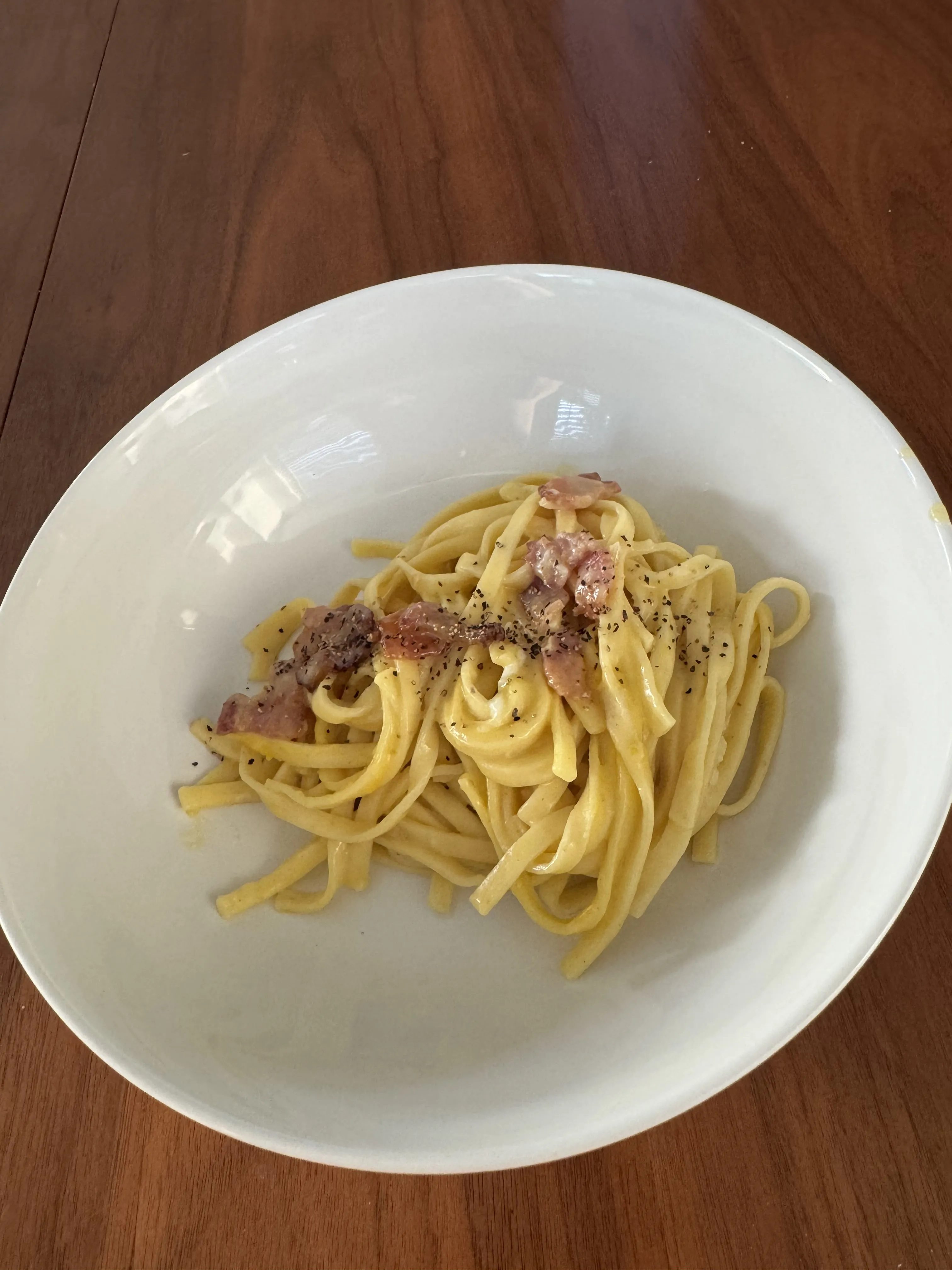 A shallow bowl of fettuccine carbonara, glossy yellow-gold with cracked pepper and small pink pieces of guanciale scattered through.