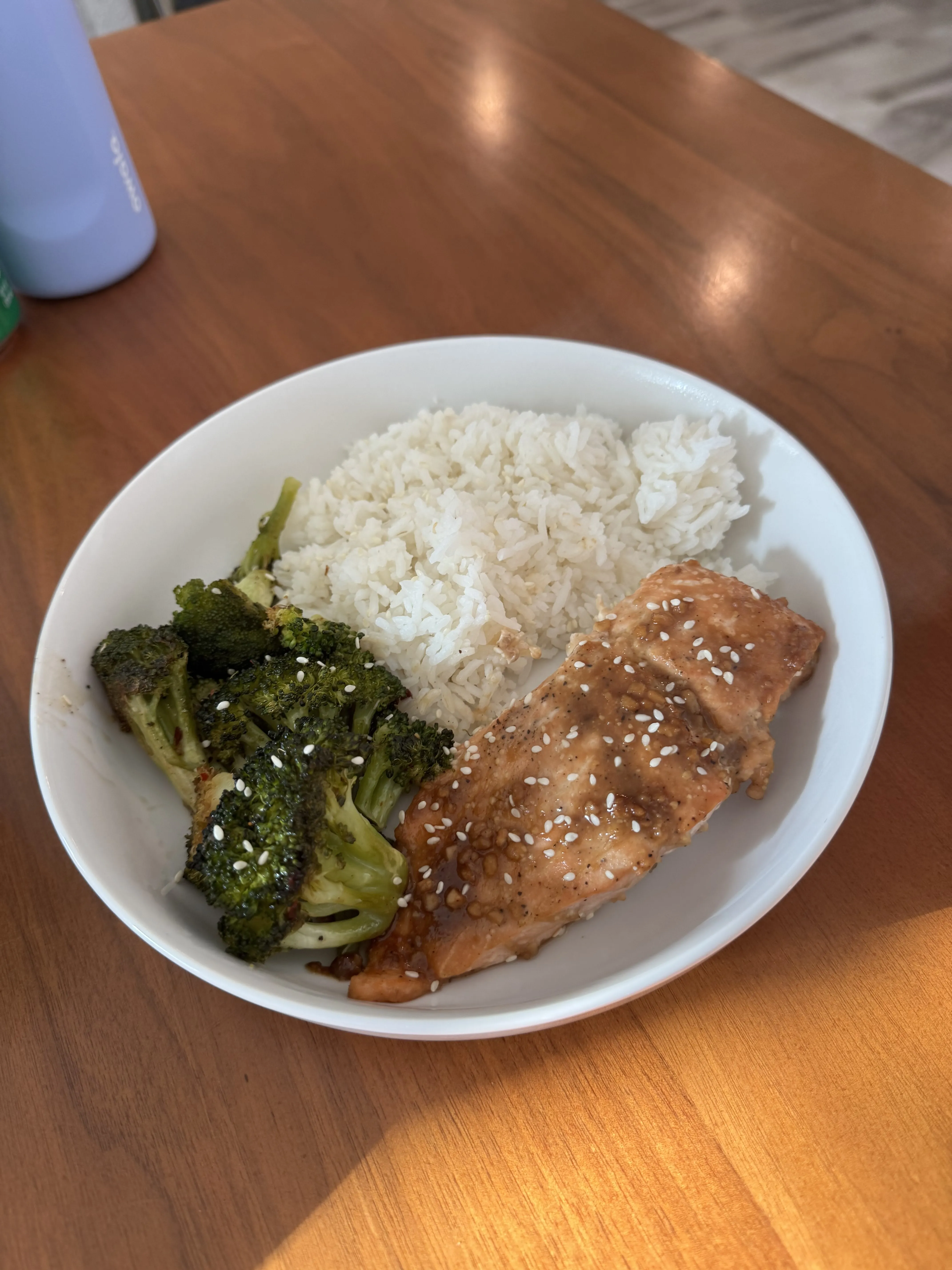 A white bowl with white rice, pieces of teriyaki chicken shining with sauce and sesame seeds, and charred florets of broccoli.