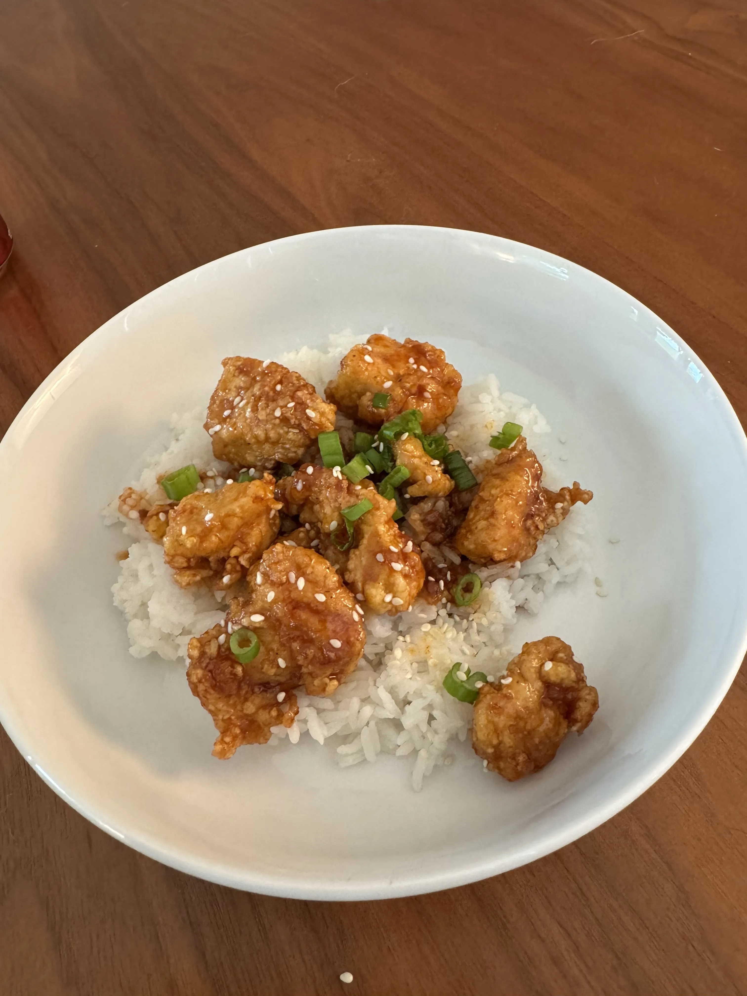 A bowl of rice topped with sesame-glazed crispy chicken and a scatter of sliced green onions.