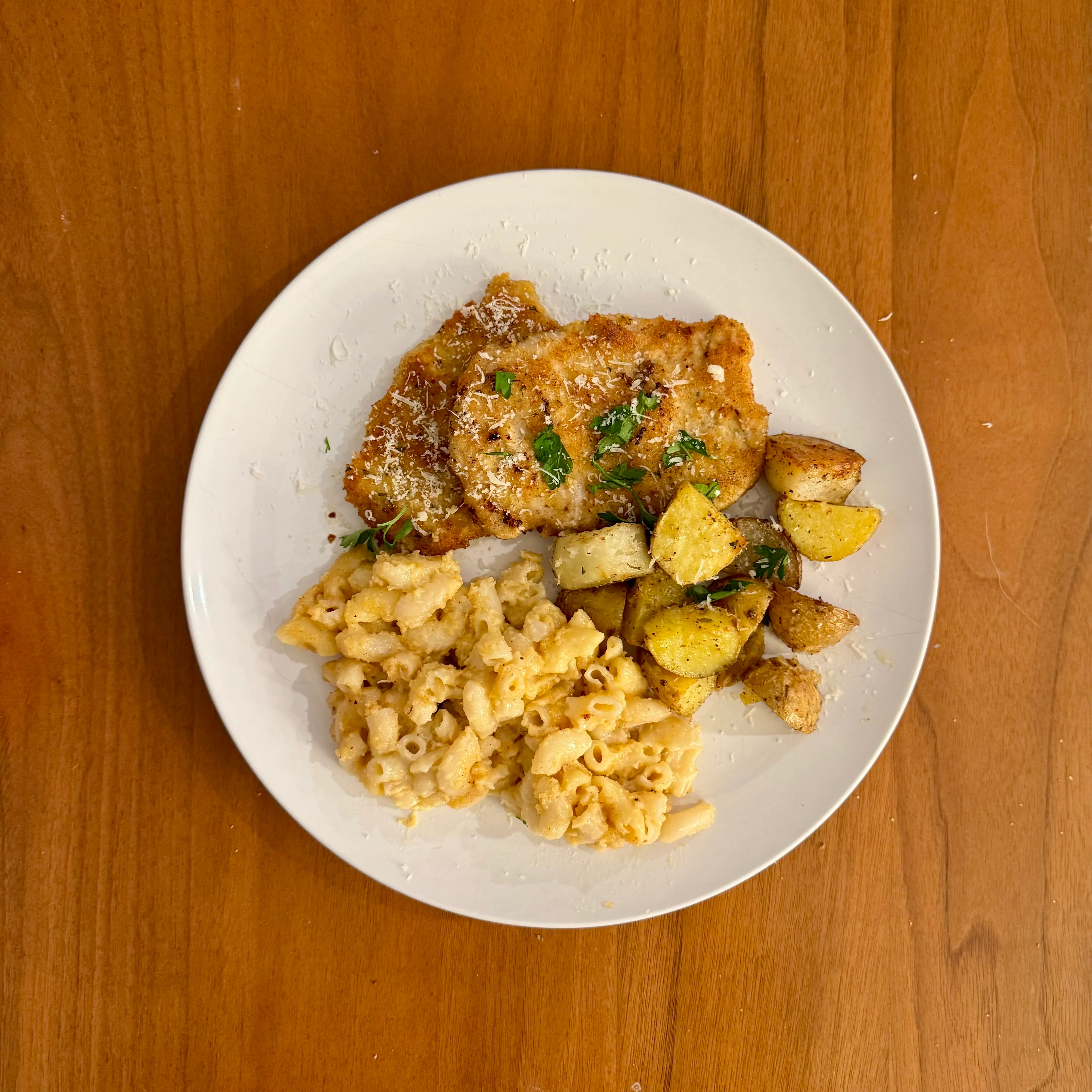 A plate of chicken parmesan with melted cheese and parsley, a side of small-shell mac and cheese, and golden roasted potato chunks.