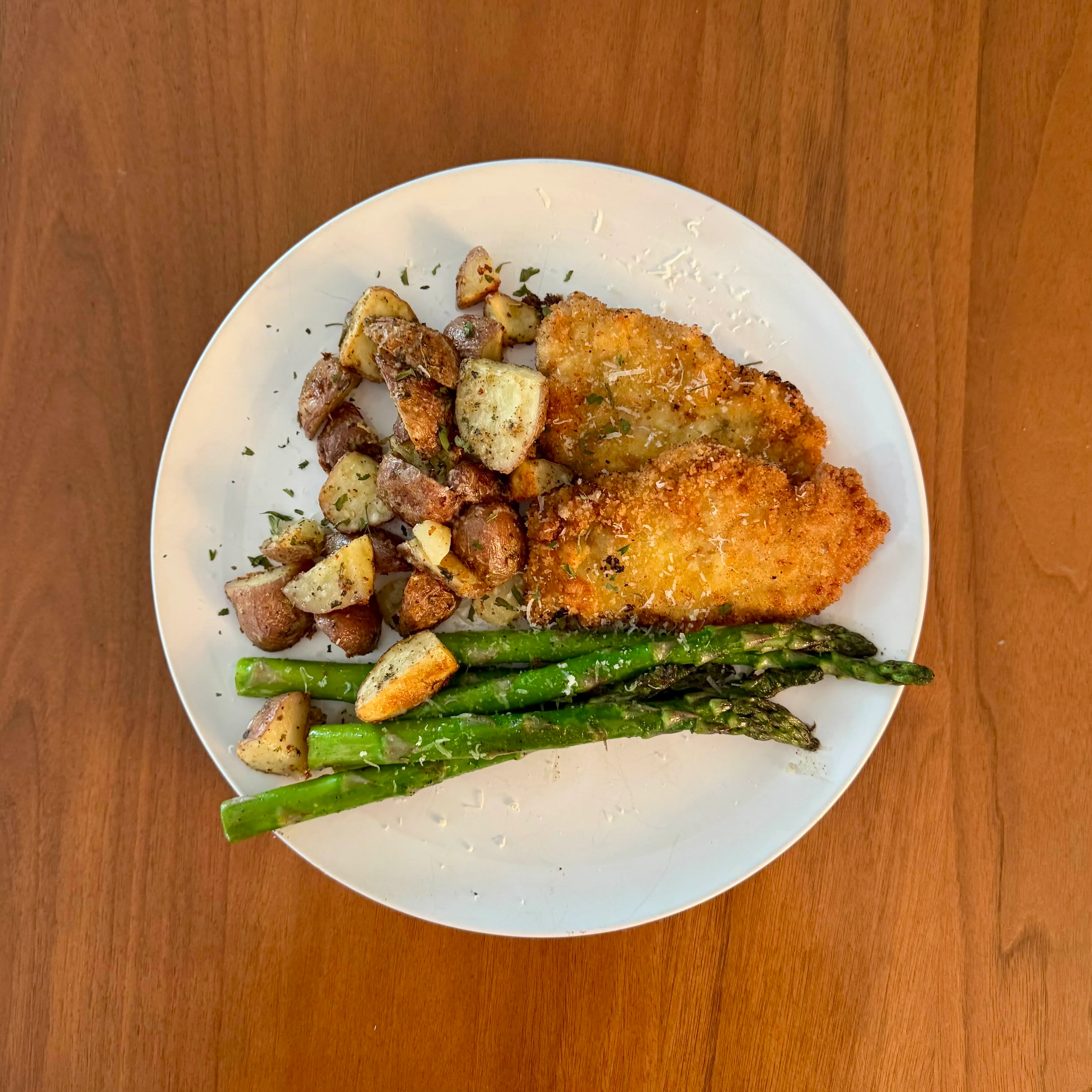 A plate of breaded chicken cutlet with a crisp golden crust, next to roasted potato cubes and a few long asparagus spears, dusted with parmesan.