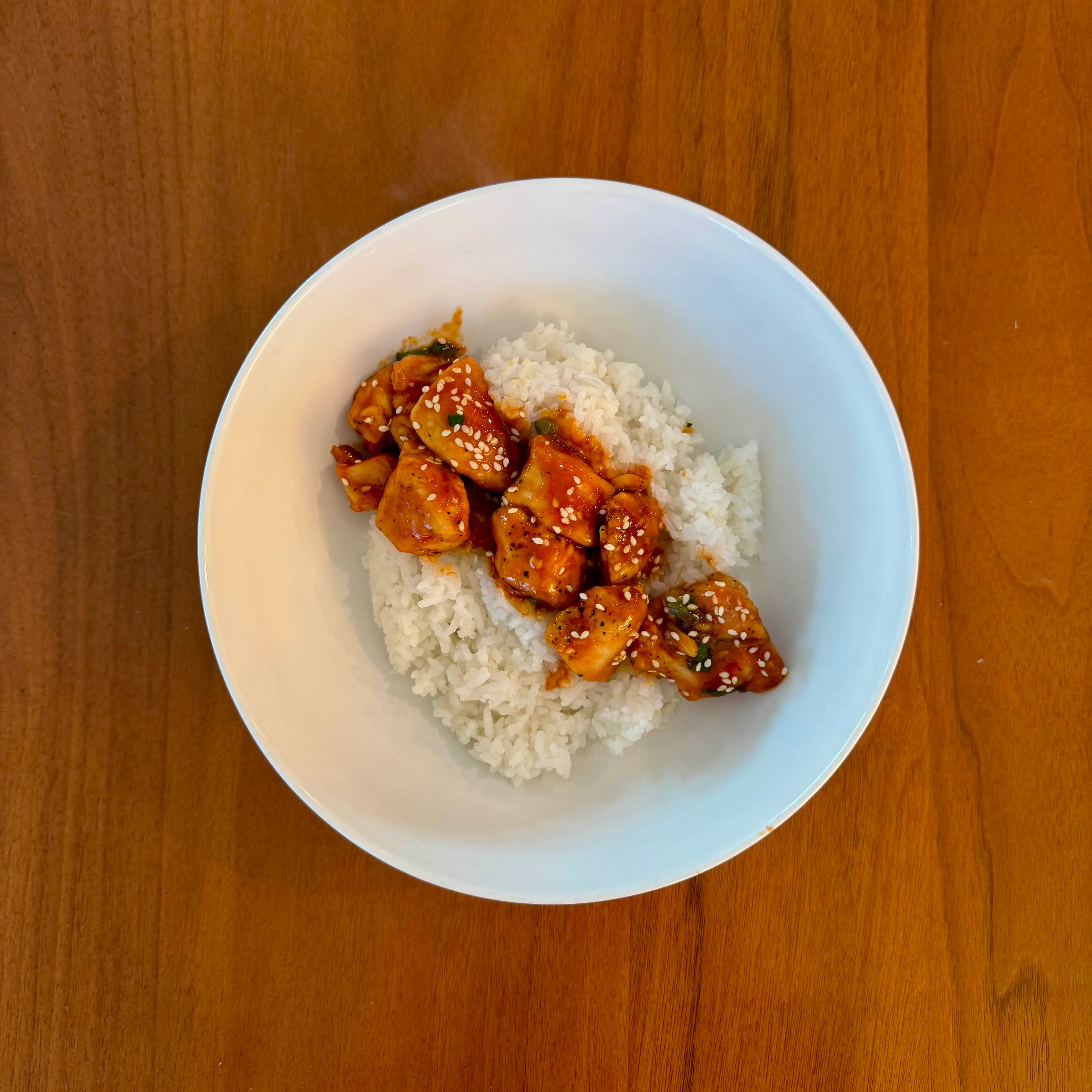 A bowl of white rice with craggy pieces of orange chicken in a dark glossy sauce, dusted with sesame seeds.