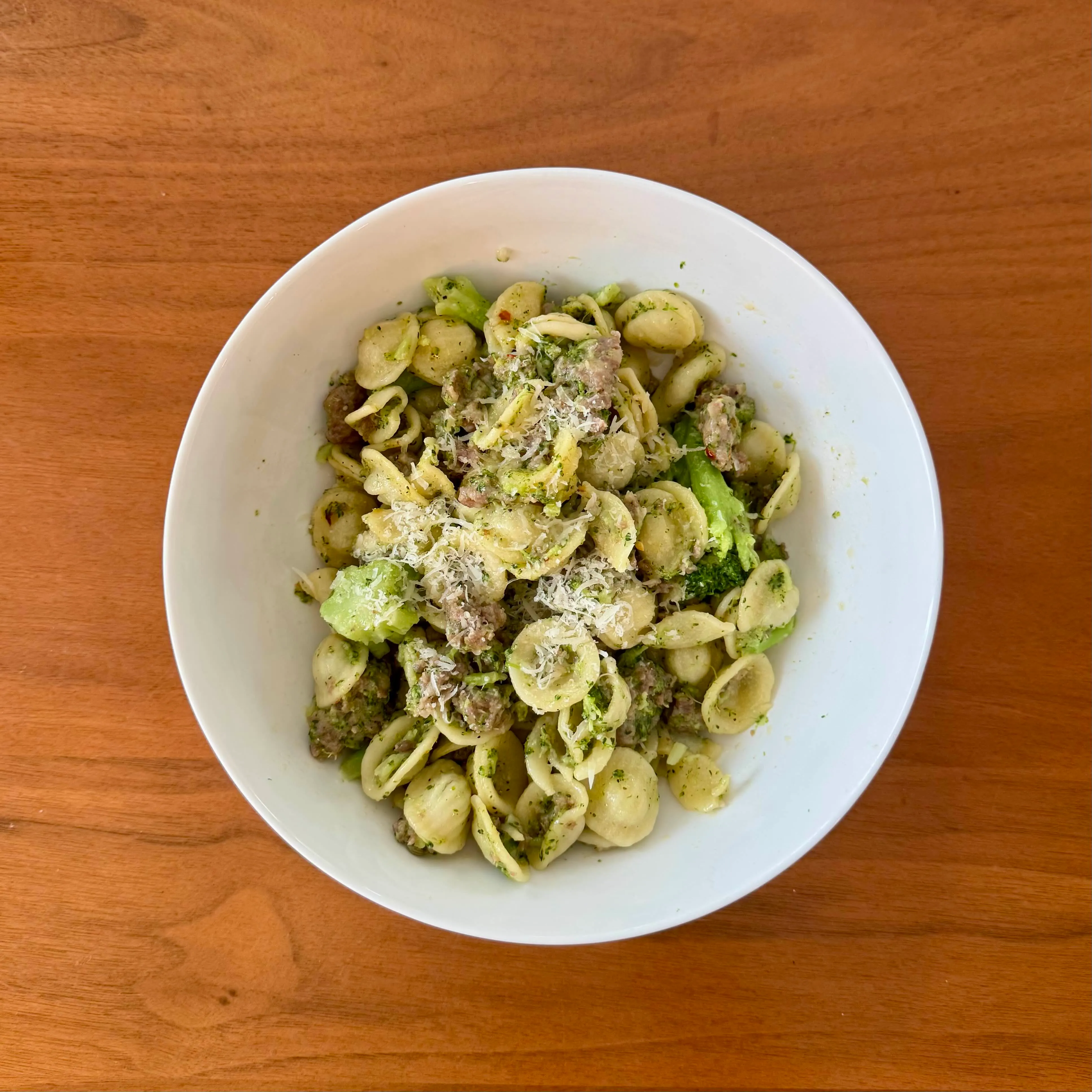 A white bowl of orecchiette tossed with crumbled Italian sausage, bright green broccoli florets, and shaved parmesan.