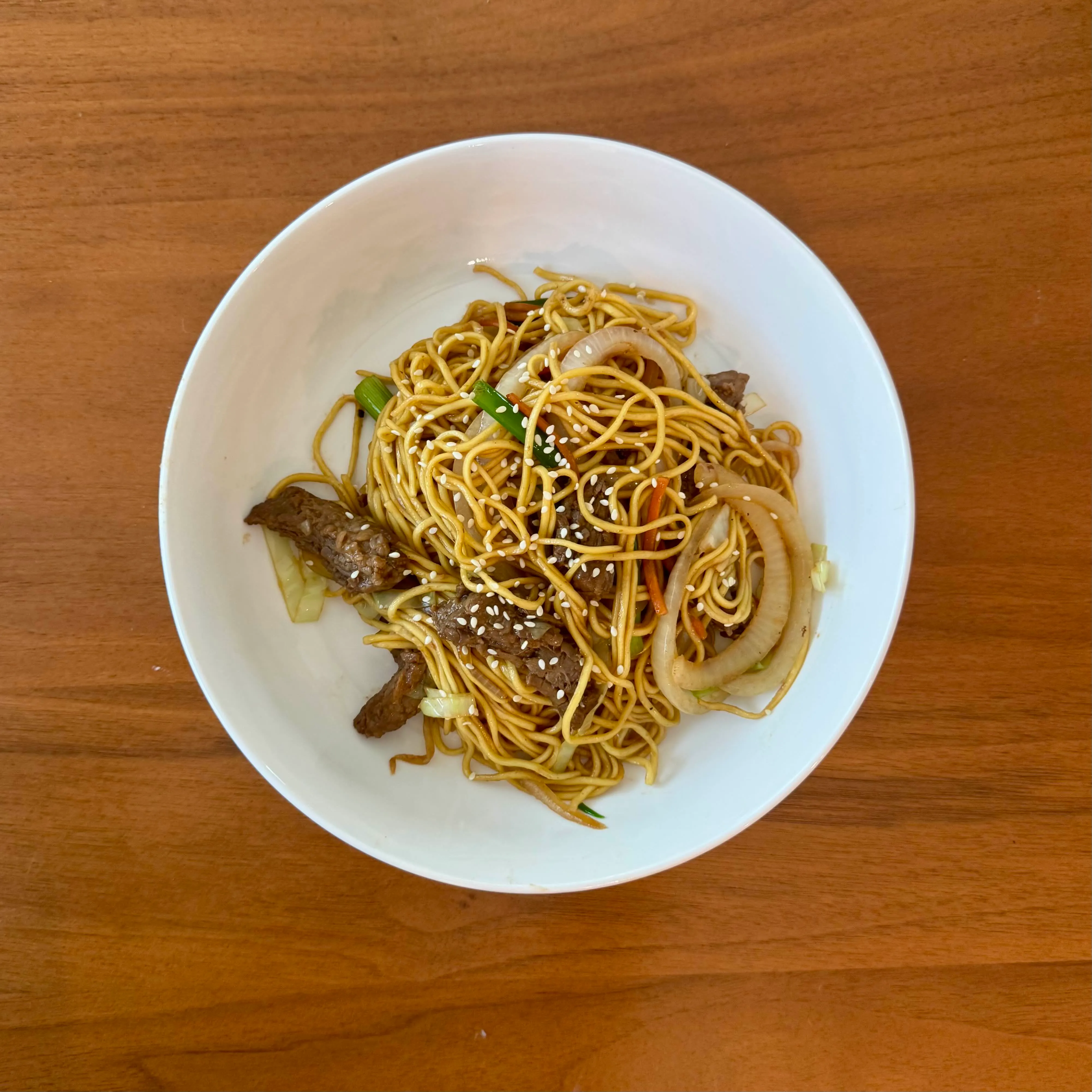 A deep bowl of beef lo mein - thin noodles tangled with strips of beef, sliced onions, matchstick carrots, and sesame seeds.