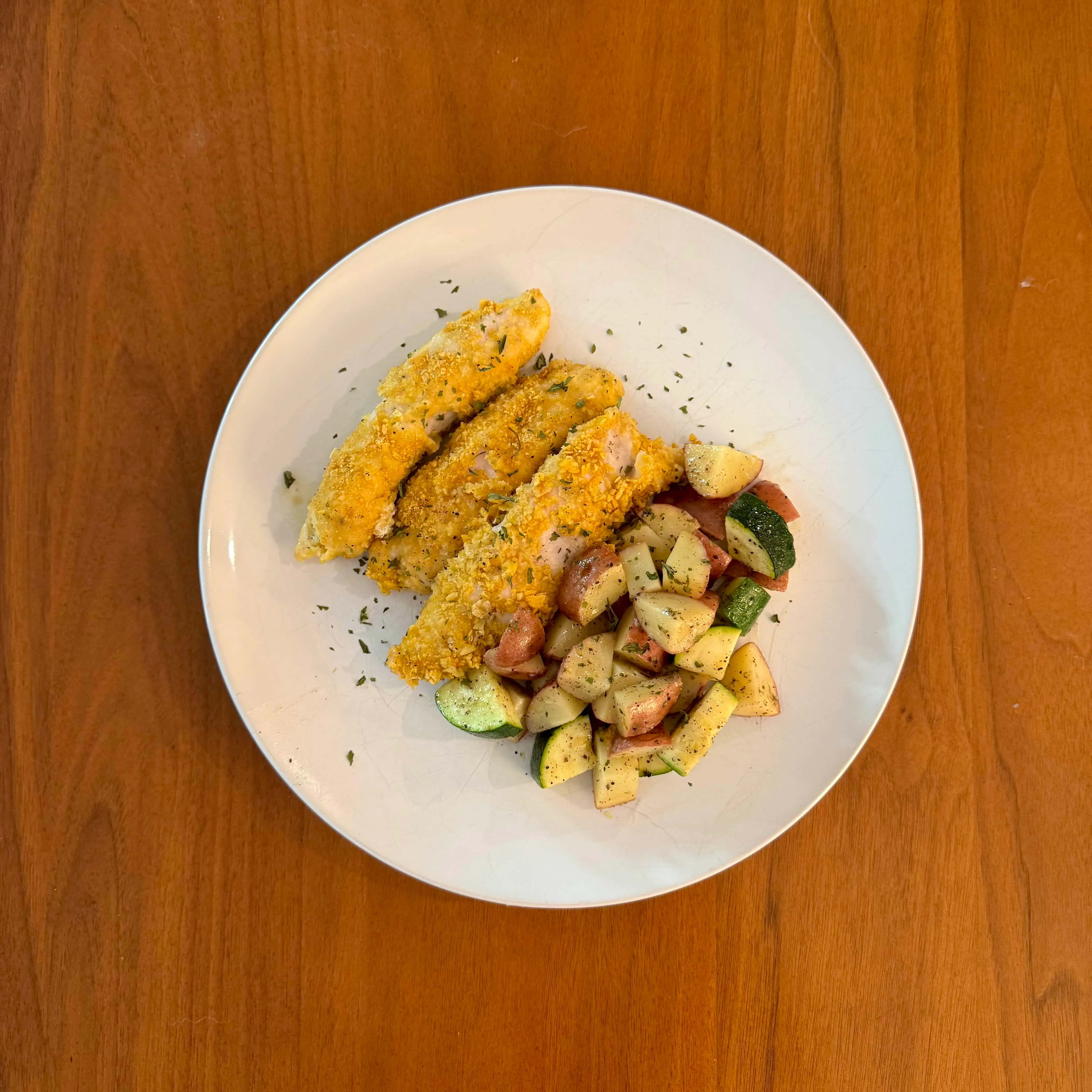 Three golden-crusted chicken tenders beside a summer vegetable side of zucchini, yellow squash, and red potato cubes.