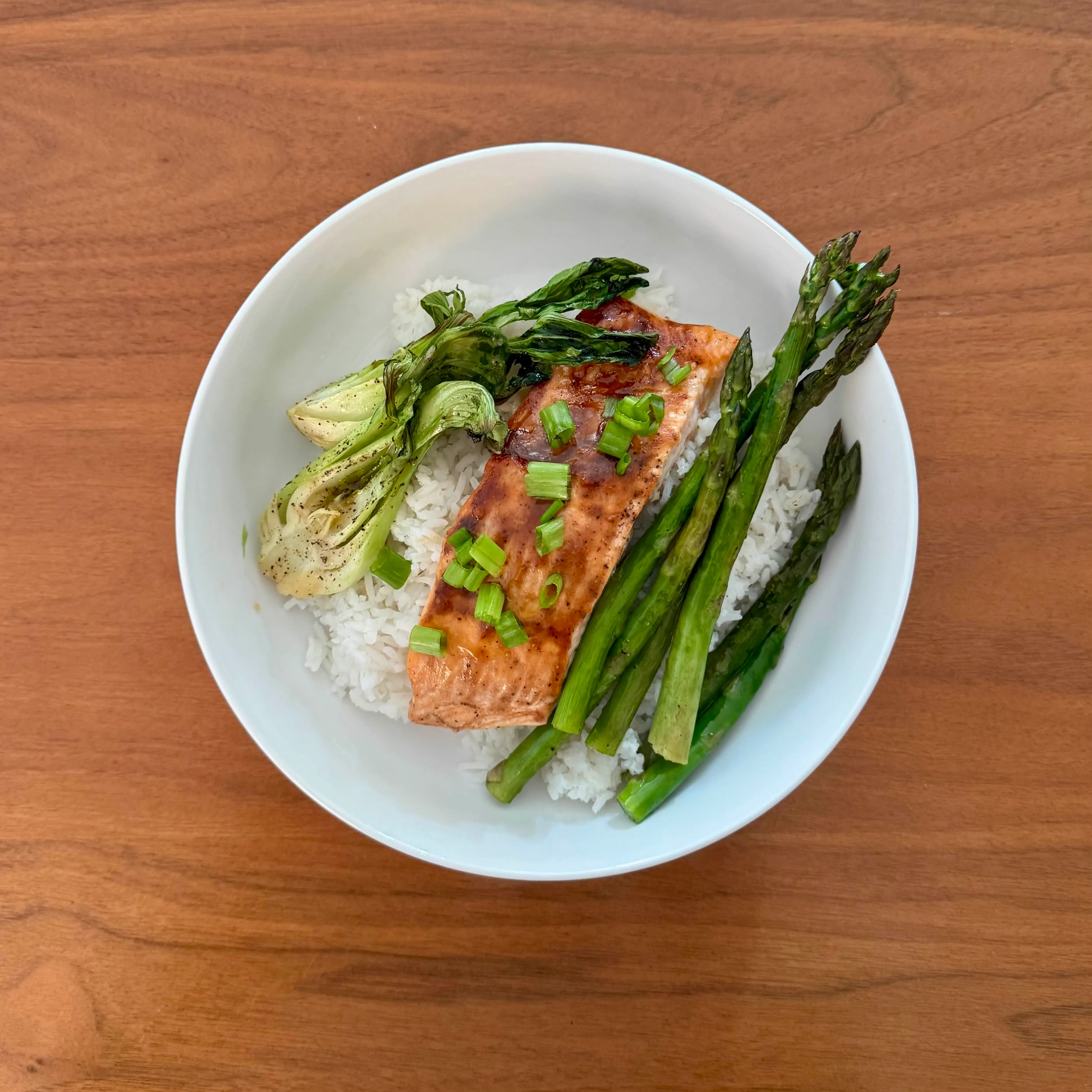 A bowl of white rice with a glazed salmon fillet sprinkled with scallions, charred bok choy halves and long asparagus spears on the side.
