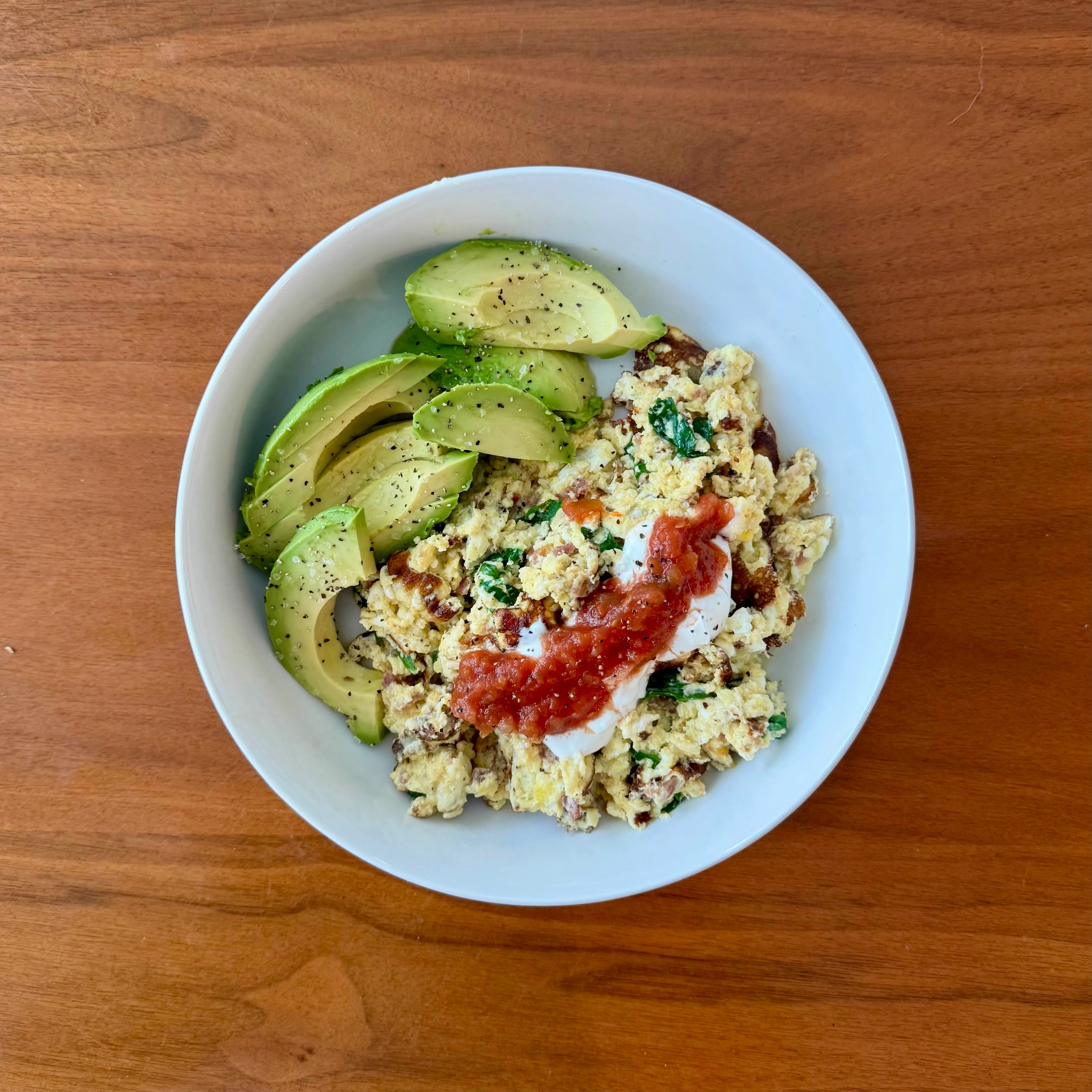 A breakfast bowl with scrambled eggs and greens, a dollop of sour cream and salsa on top, and fanned slices of avocado along one side.