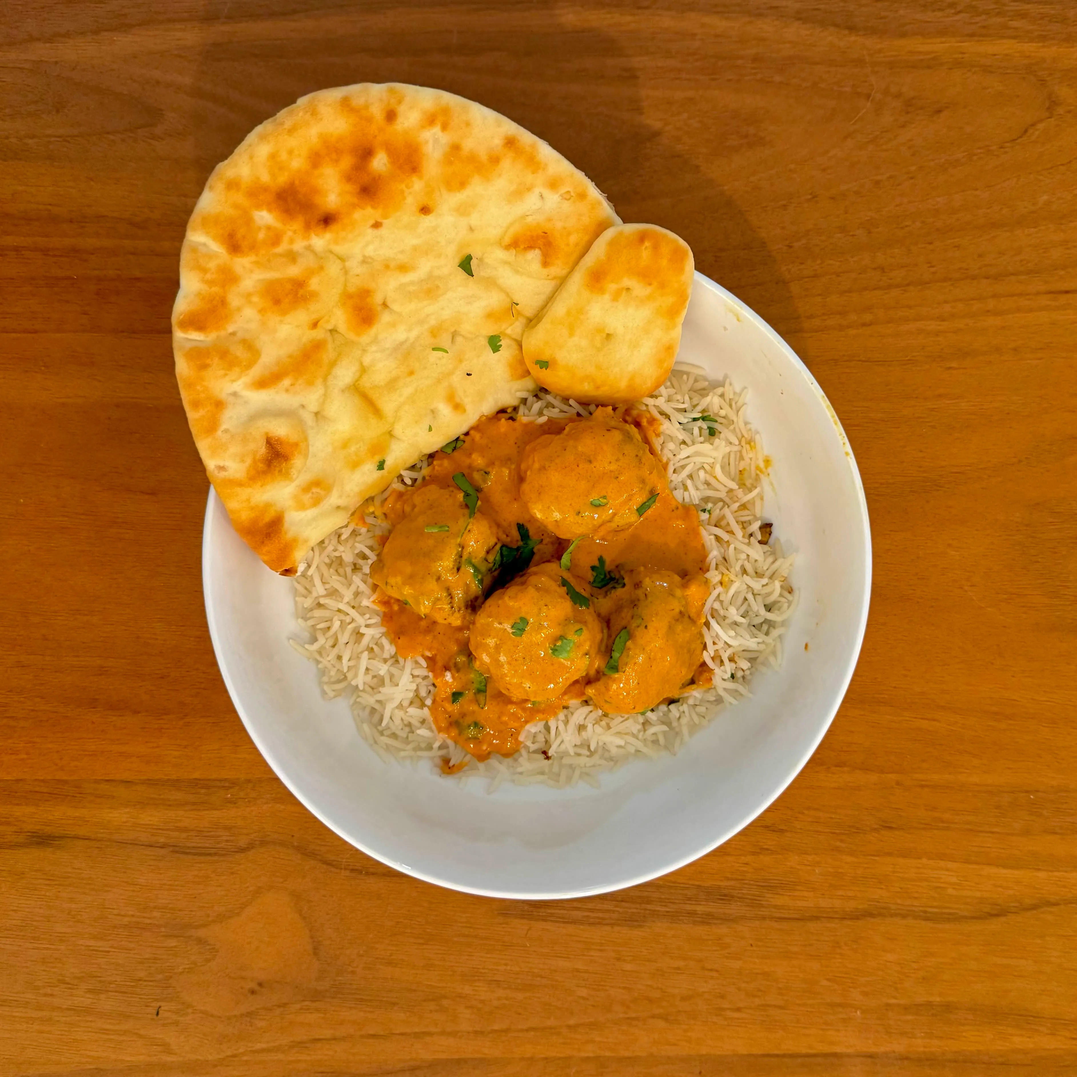 A bowl of basmati rice with meatballs in a marigold-orange curry, plus a half-round of naan tucked on top.