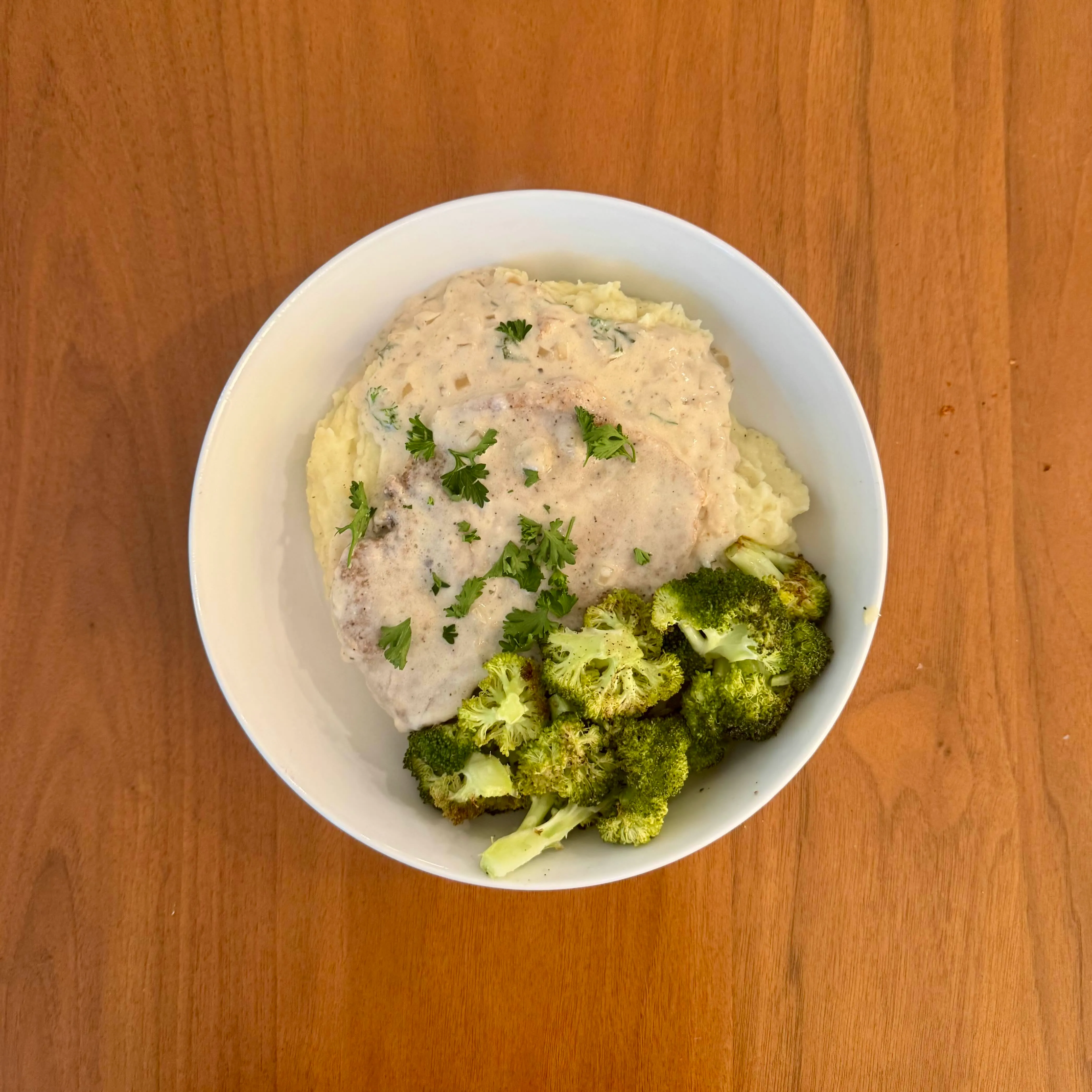 A mound of mashed potatoes with a chicken breast in a pale cream sauce and roasted broccoli florets on the side, parsley scattered over.