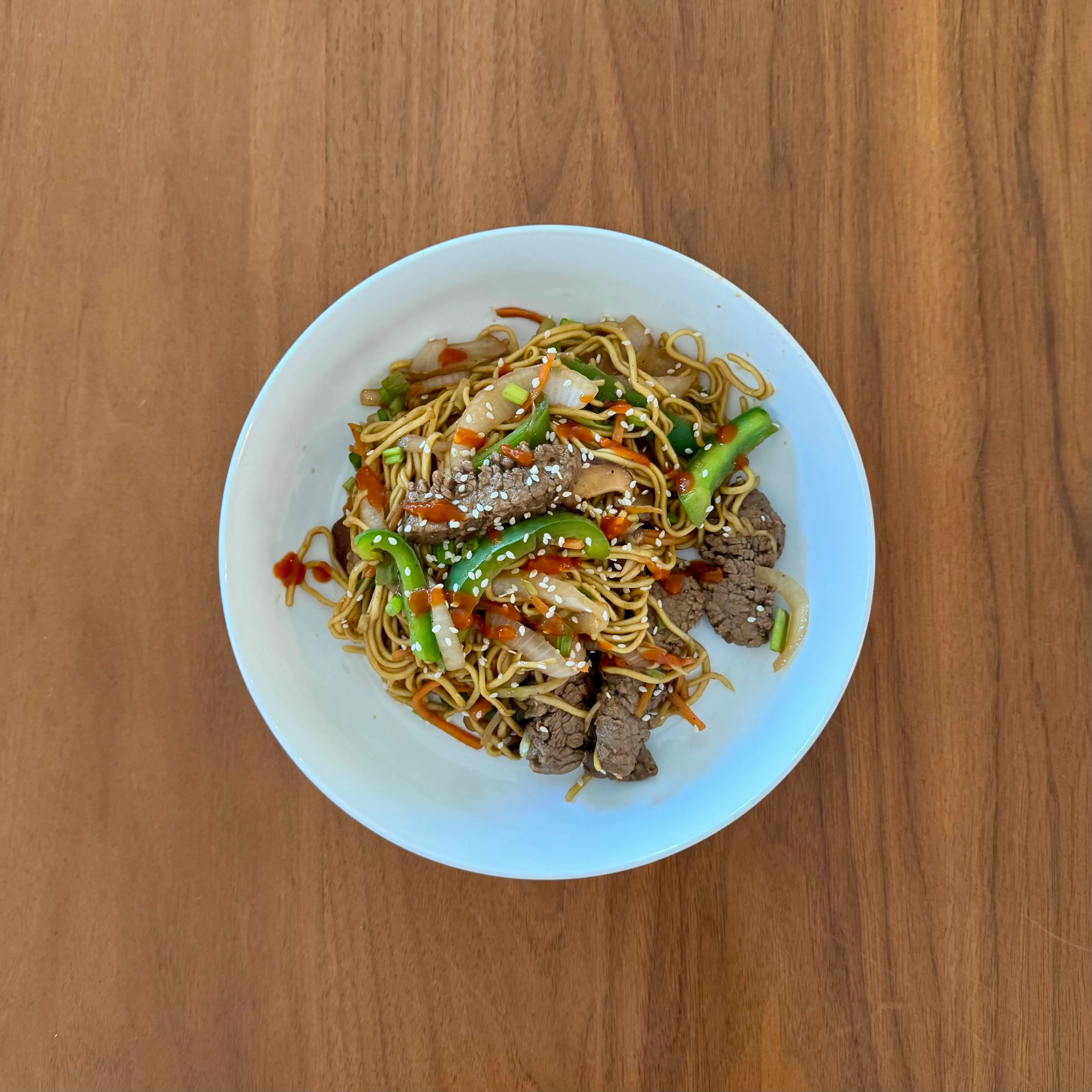 A bowl of beef lo mein tangled with green beans and matchstick carrots, dotted with sesame seeds and a drizzle of chili oil.
