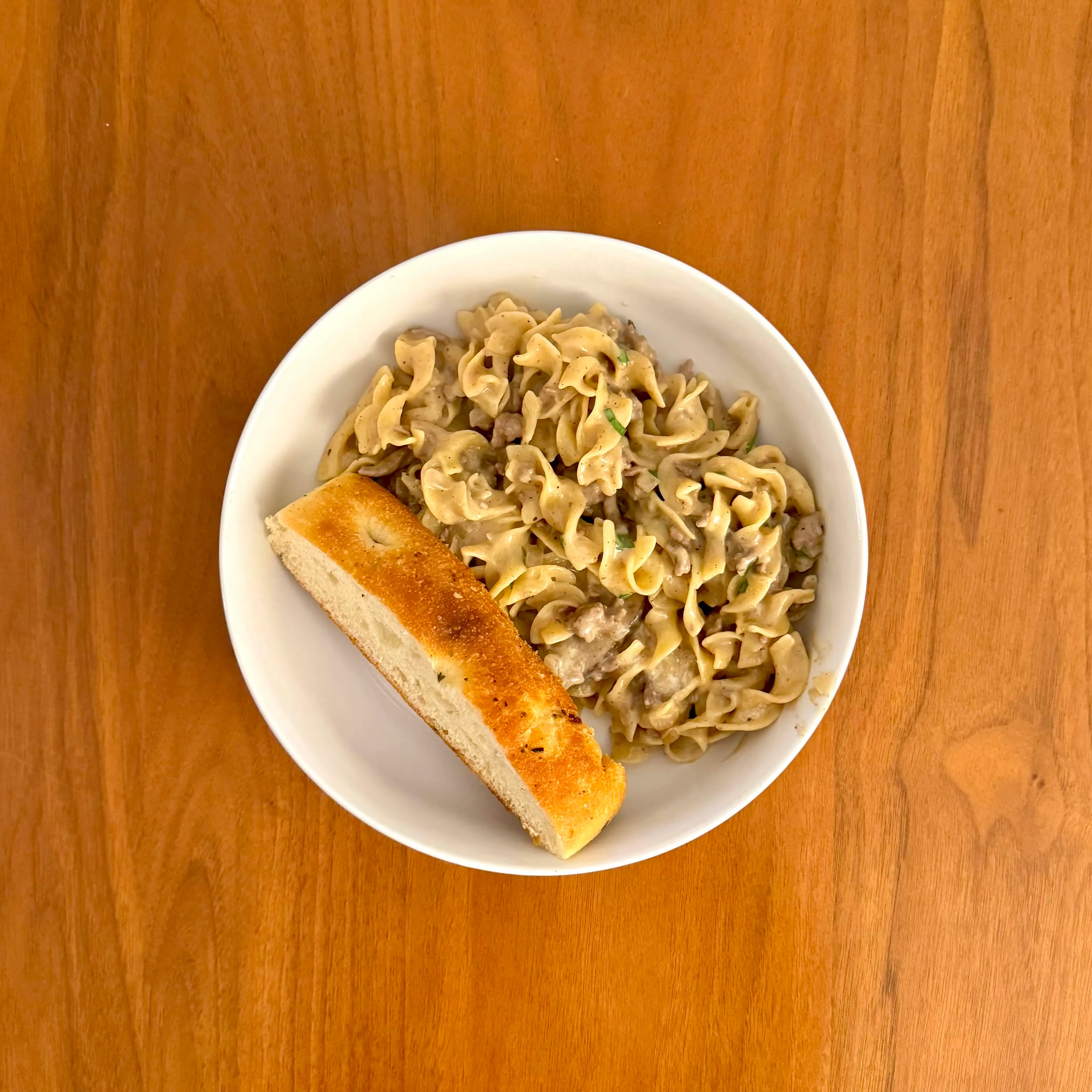 A white bowl of creamy beef stroganoff over wide egg noodles, with a thick slice of toasted bread tucked along the side.