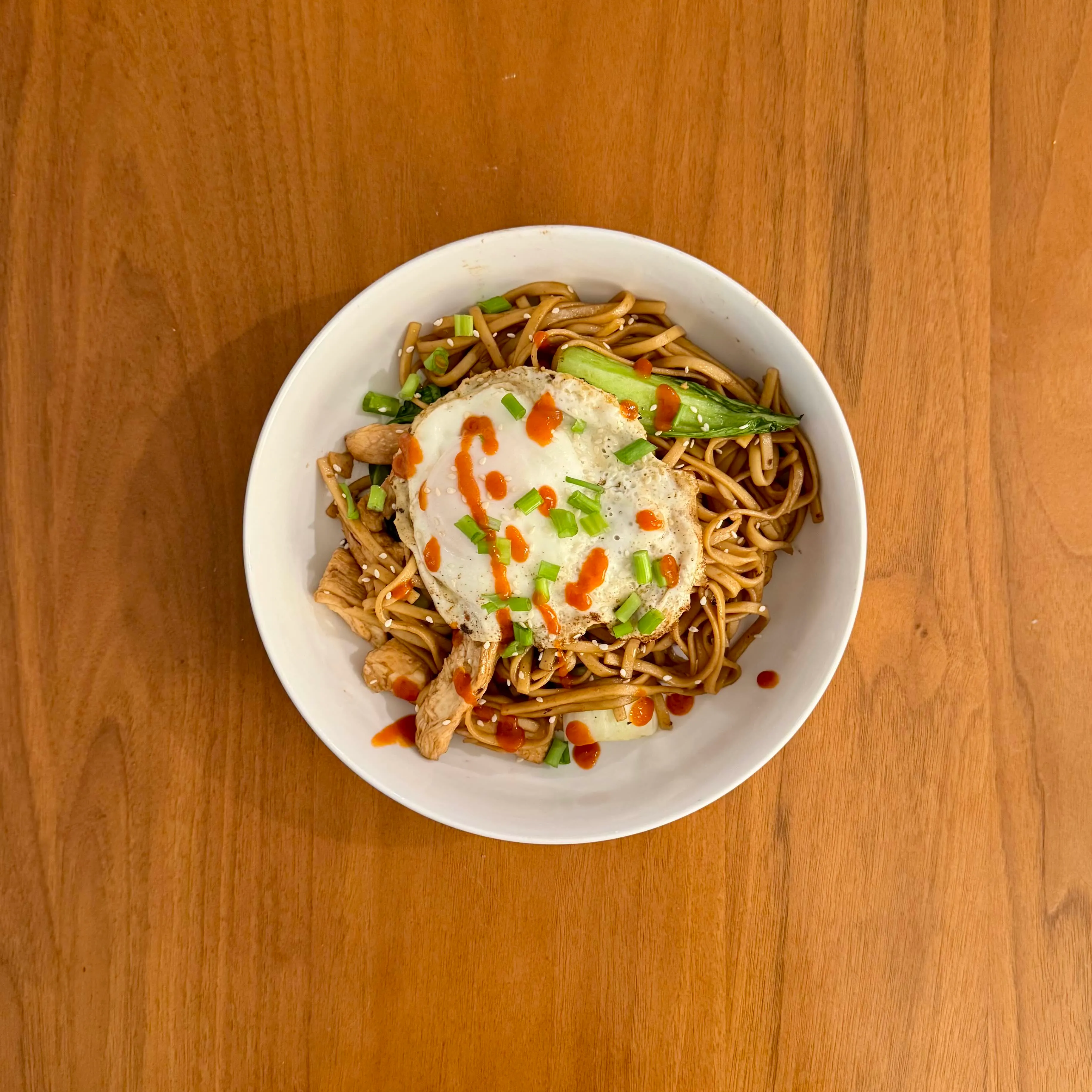 A bowl of soy-garlic noodles with a fried egg and a bok choy half on top, bright orange sriracha striped across.