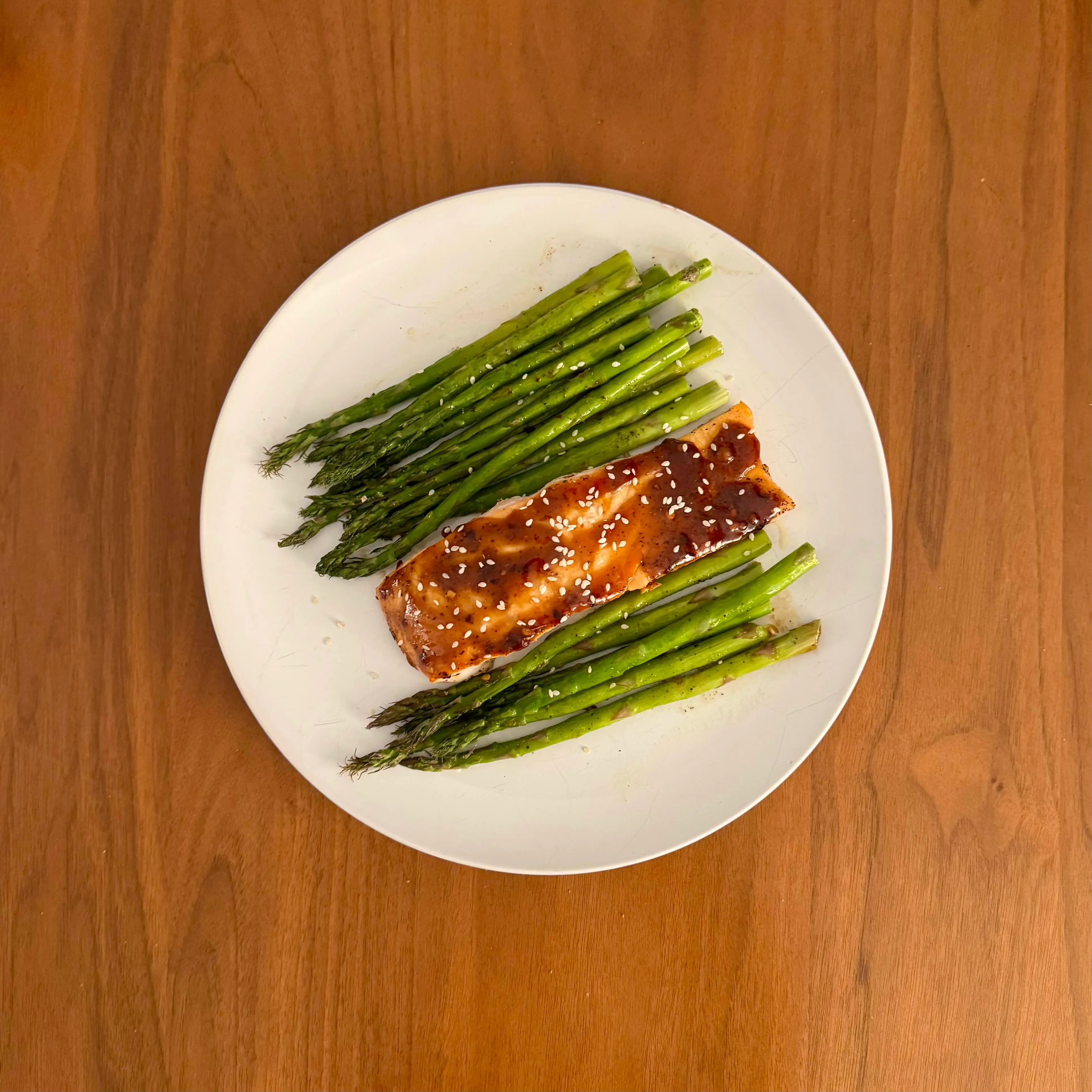A glazed salmon fillet on a plate surrounded by long spears of asparagus.