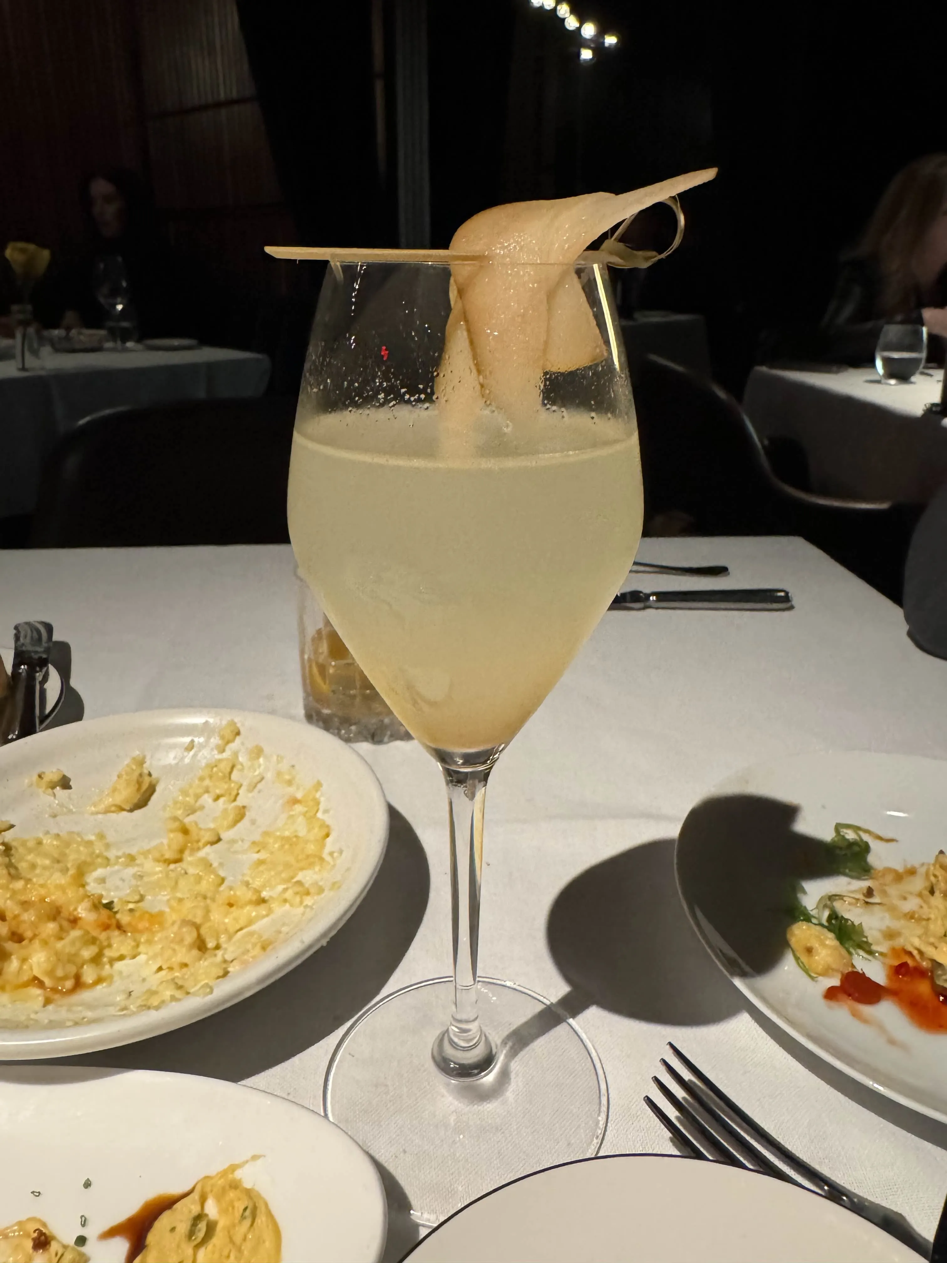 A champagne flute of pale yellow cocktail with a curled paper-thin tuile garnish balanced across the rim, on a white-clothed table in a dim restaurant.