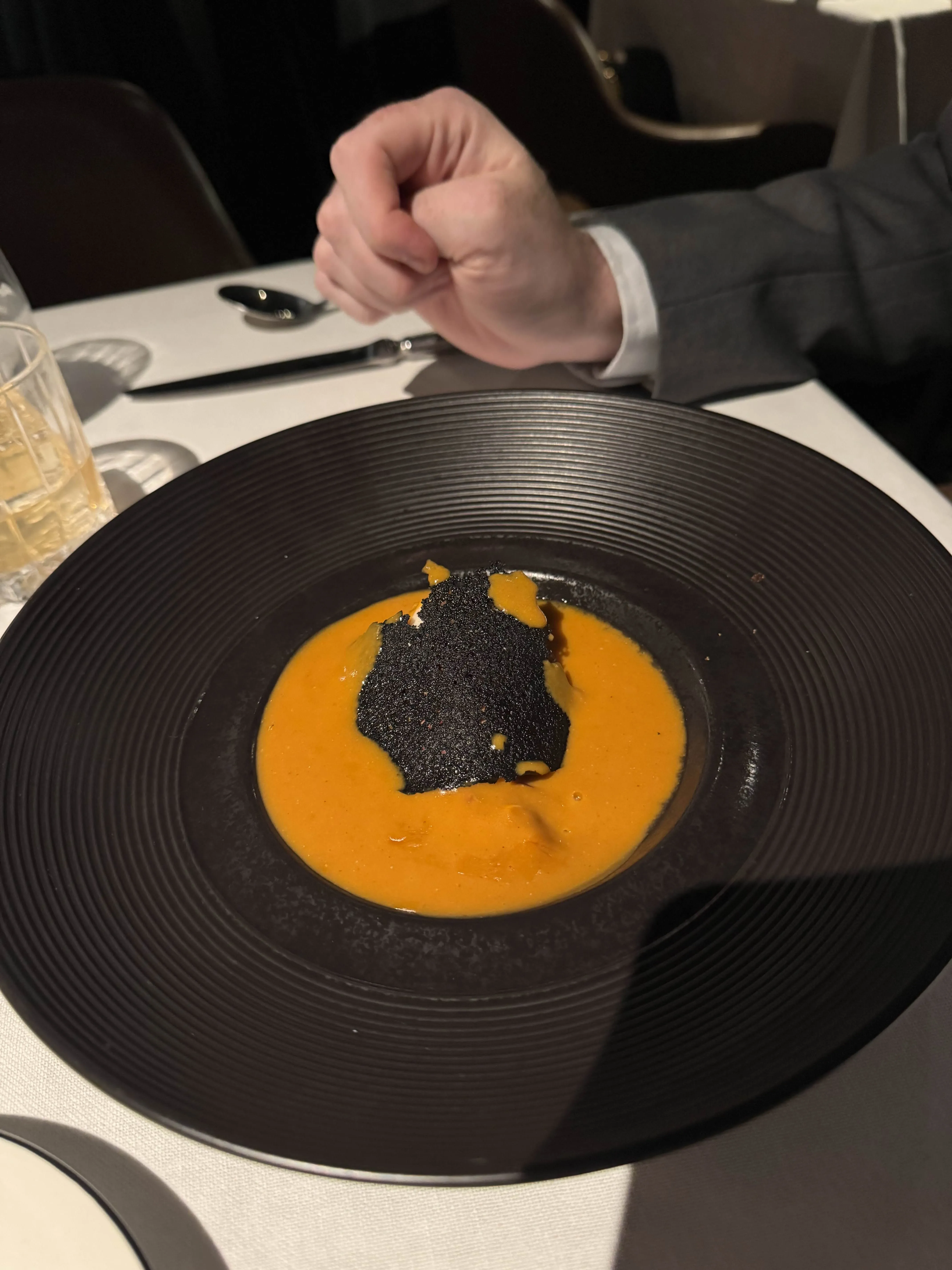 A black ridged bowl with a pool of orange bisque and a jet-black parmesan crisp floating on top, the diner's hands resting on the white table.