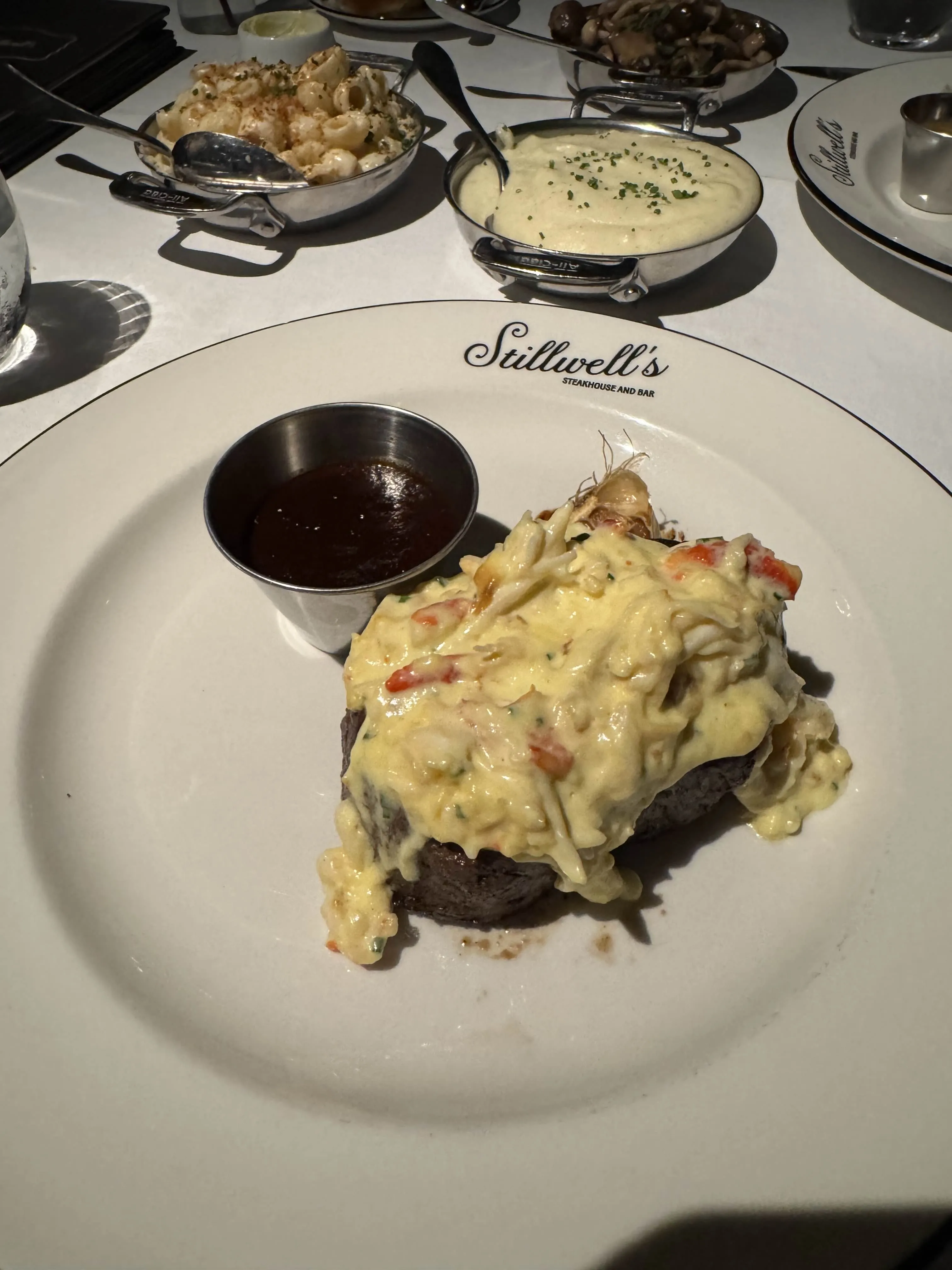 A filet mignon topped with lump crab in a hollandaise-like sauce on a plate printed with "Stillwell's Steakhouse and Bar," with a small ramekin of bordelaise and the sides plated behind.