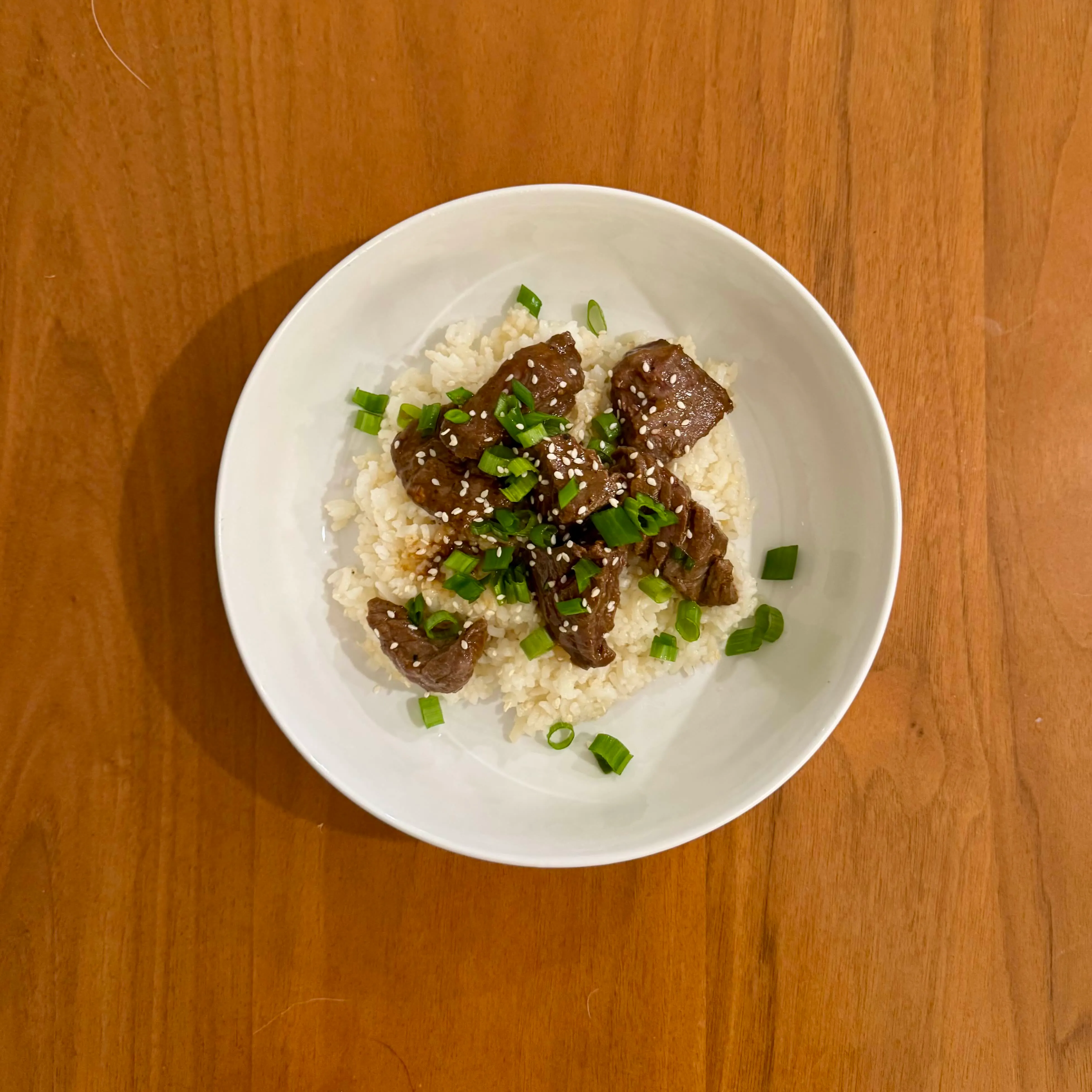 A bowl of white rice with cubes of glossy dark-glazed beef, sprinkled with sesame seeds and sliced scallions.