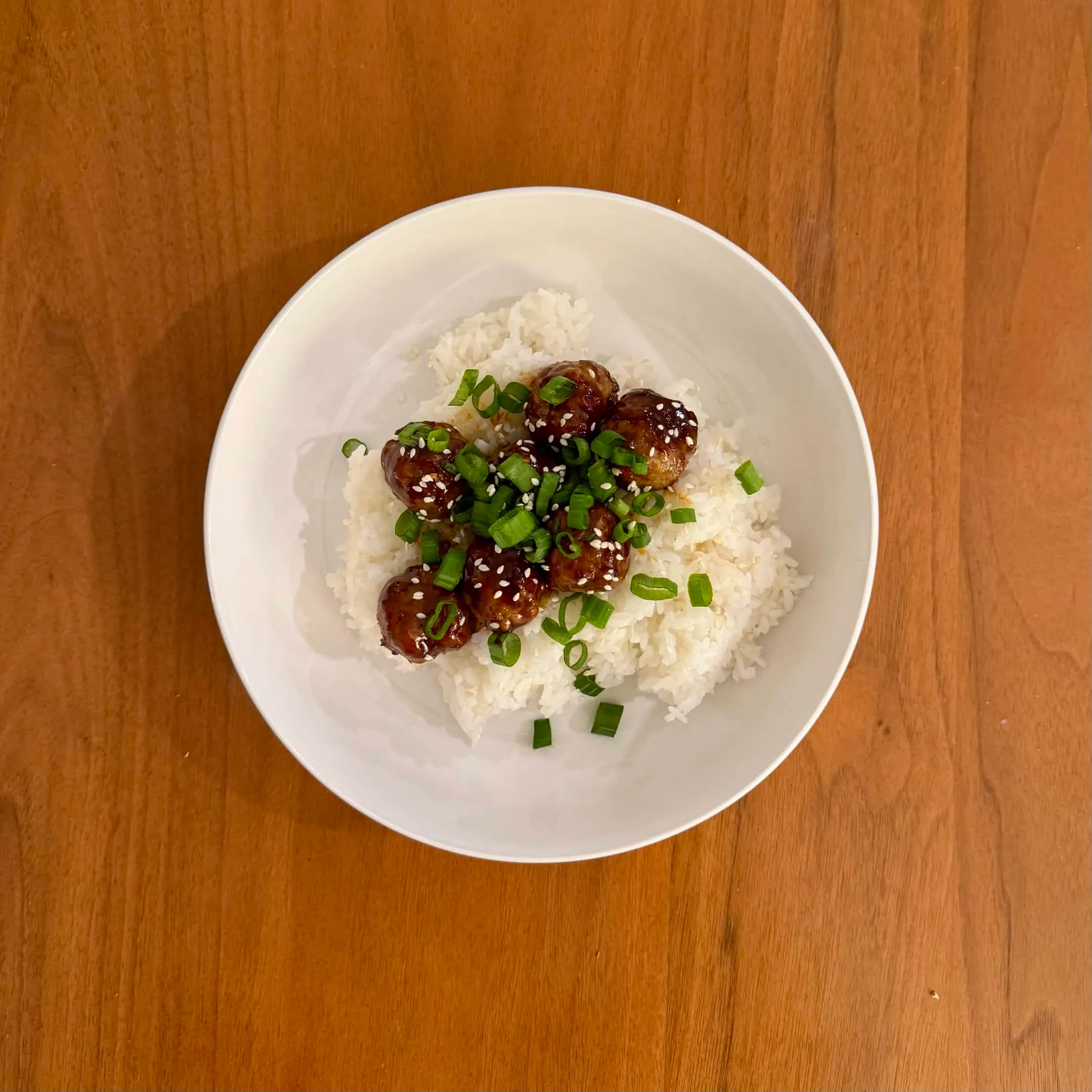 A shallow bowl of white rice with teriyaki meatballs glazed dark brown, scattered with sesame seeds and sliced green onions.