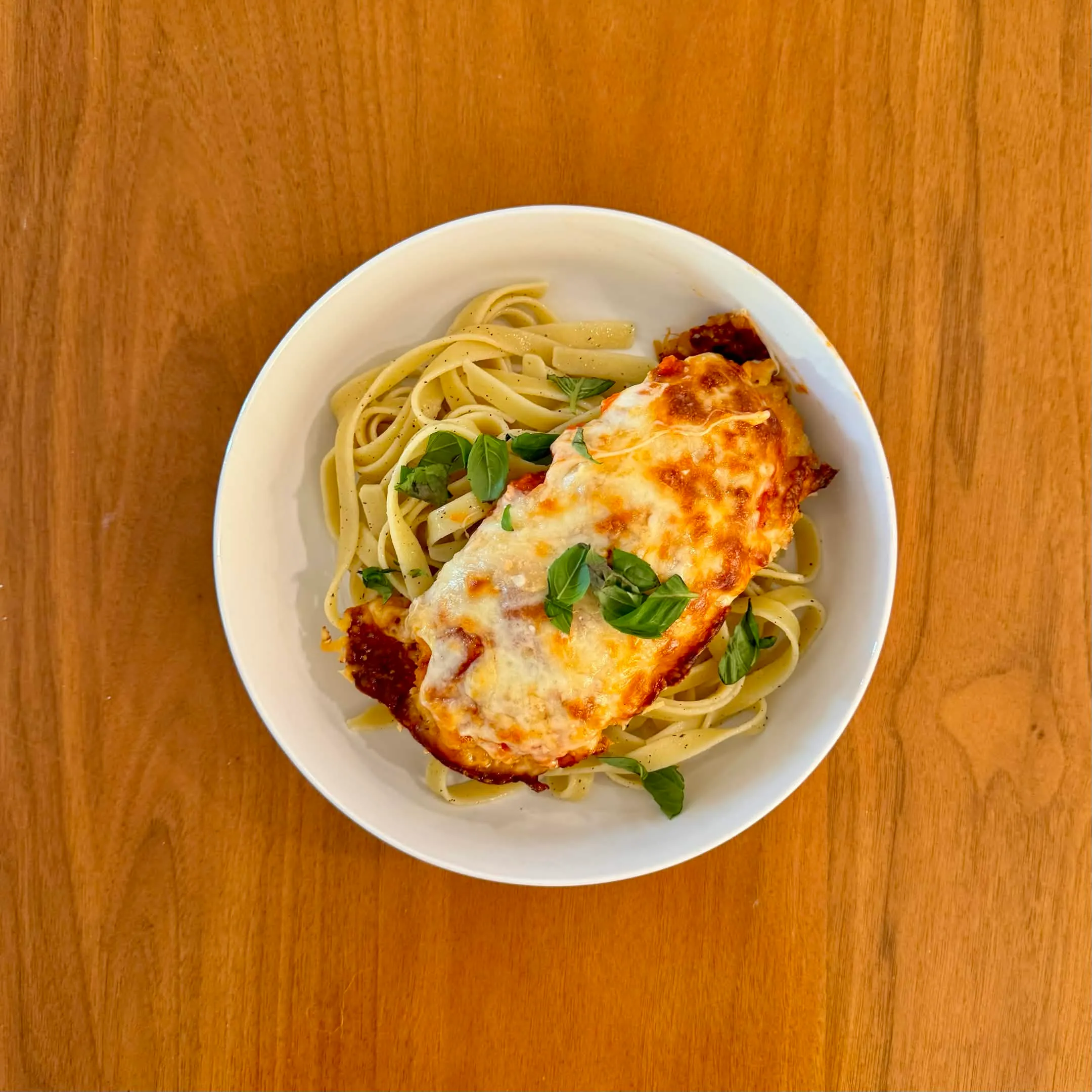 A bowl of fettuccine tossed in red sauce with a piece of breaded chicken topped with melted mozzarella and torn basil.
