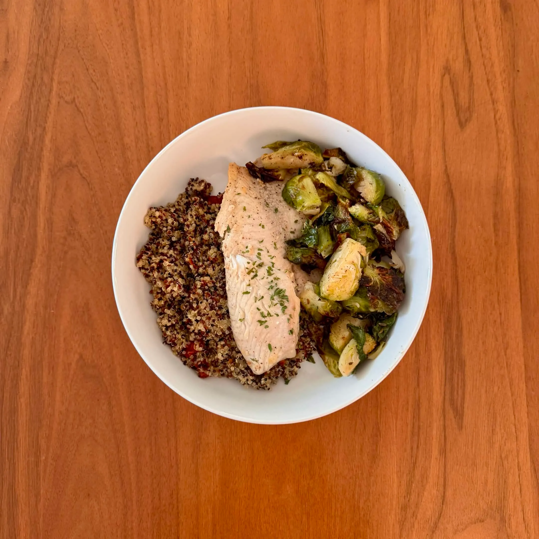 A bowl with a lemon-herb chicken breast over a tri-colored quinoa pilaf and a pile of roasted halved brussels sprouts.