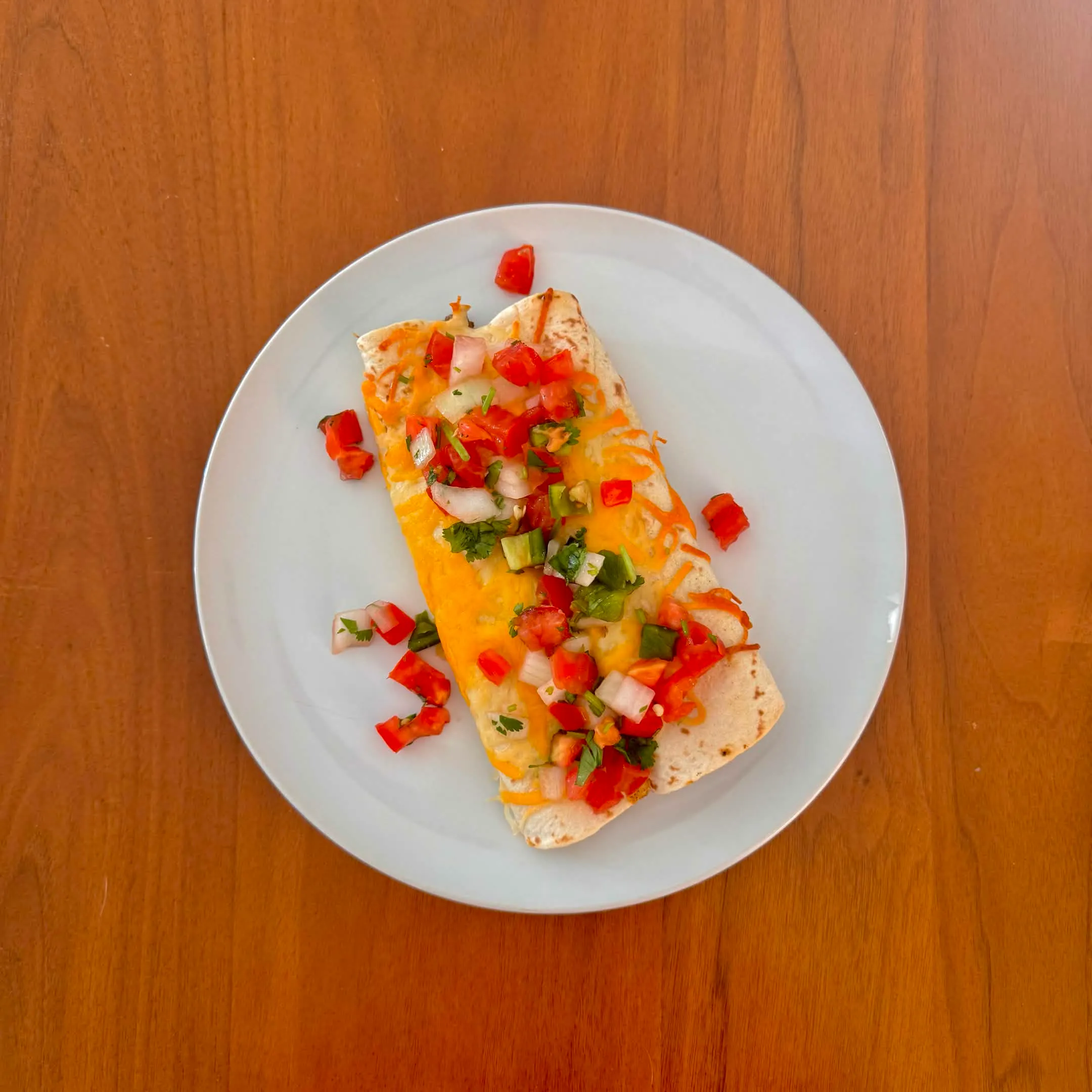 A plate with a single cheese-topped rolled enchilada covered in pico de gallo and chopped scallions.